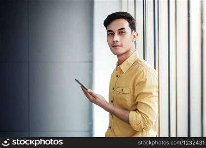 Portrait of Happy Businessman Standing by the Window in Office. Using Smartphone and Smiling. Looking at Camera