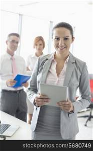 Portrait of happy businesswoman holding digital tablet with colleagues in background at office