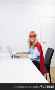 Portrait of happy businesswoman in superhero costume using laptop at office