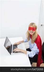 Portrait of happy businesswoman in superhero costume working on laptop in office