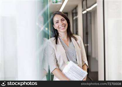 Portrait of happy businesswoman standing with documents by wall at office