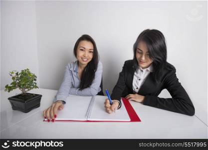Portrait of happy businesswoman with colleague writing in spiral notebook at office desk