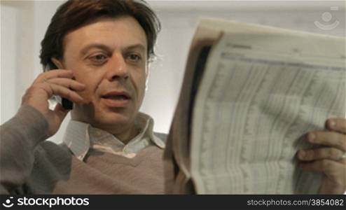 Portrait of happy caucasian adult man sitting on sofa at home, reading stock exchange listings on newspaper and talking on cordless telephone with trader