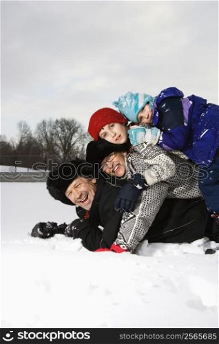 Portrait of happy Caucasian family of four lying stacked on top of each other in snow smiling at viewer.