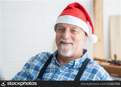 Portrait of happy Caucasian senior carpenter kind man with white beard and mustache wearing santa hat smiling with handmade wooden work on background at workshop on Christmas holiday