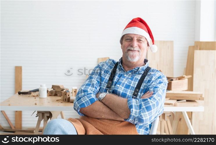 Portrait of happy Caucasian senior carpenter man with beard and mustache wearing santa hat smiling sitting with arms crossed with handmade wooden work on background at workshop on Christmas holiday