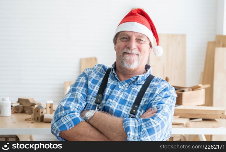 Portrait of happy Caucasian senior carpenter man with beard and mustache wearing santa hat smiling stand with arms crossed with handmade wooden work on background at workshop on Christmas holiday