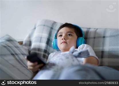 Portrait of happy child wearing headphones and looking up watching TV, Kid sitting on sofa holding remote control, Happy young boy listening to music relaxing in living room at home