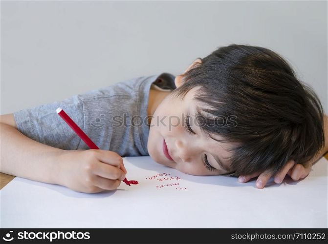 Portrait of happy child writing a messages to his mother, Preschool kid using red colour writing and drawing on white paper, Little boy lying head down on table drawing red heart for mam.