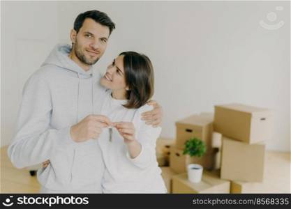 Portrait of happy couple embrace in empty room, hold keys, just arrived in own apartment, need to unpack personal belongings in boxes, look gladfully at camera, being happy owners of flat or house