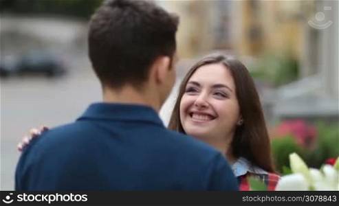 Portrait of happy couple in love hugging in the street with smiling woman&acute;s face in foreground. Beautiful happy woman with bouquet of flowers embracing her beloved boyfriend tenderly with hands and looking at camera with radiant toothy smile.