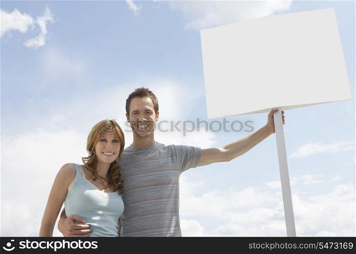 Portrait of happy couple with blank sign board against cloudy sky