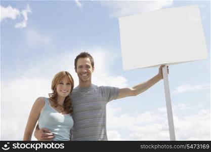 Portrait of happy couple with blank sign board against cloudy sky