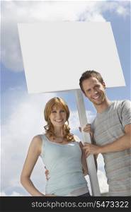 Portrait of happy couple with blank sign over cloudy sky