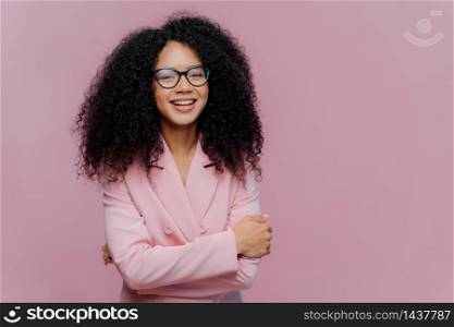 Portrait of happy curly haired businesswoman wears formal jacket, laughs positively, enjoys successful business meeting, has optical glasses, isolated over purple background blank space for your promo