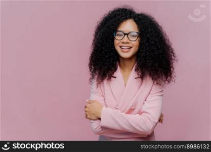Portrait of happy curly haired businesswoman wears formal jacket, laughs positively, enjoys successful business meeting, has optical glasses, isolated over purple background blank space for your promo