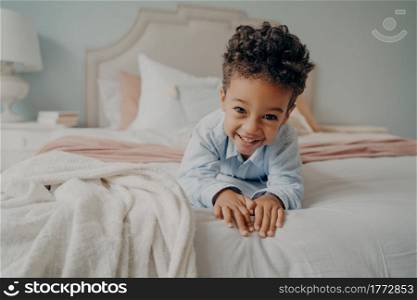Portrait of happy curly mulatto boy smiling at camera while laying on cozy bed, relaxing after fun playing session at home, adorable preschool child enjoying indoor activities and leisure time. Happy curly afro american boy laying on bed and smiling at camera