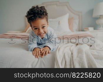 Portrait of happy curly mulatto boy smiling at camera while laying on cozy bed, relaxing after fun playing session at home, adorable preschool child enjoying indoor activities and leisure time. Happy curly afro american boy laying on bed and smiling at camera