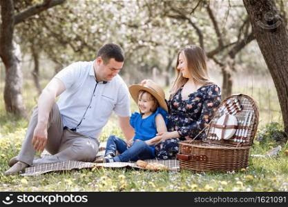 Portrait of happy family on picnic. pregnant Mom, dad and daughter sitting in spring blooming park. Young family spending time together on vacation, outdoors. Portrait of happy family on picnic. pregnant Mom, dad and daughter sitting in spring blooming park. Young family spending time together on vacation, outdoors.
