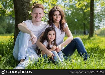 Portrait of happy family sitting on grass under a tree in park on a sunny day. Portrait of happy family in park