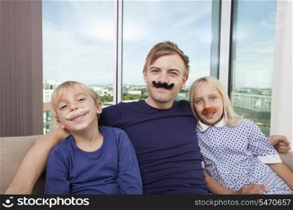 Portrait of happy father and children with artificial mustache sitting on sofa at home