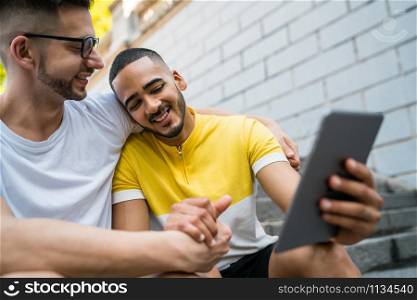 Portrait of happy gay couple spending time together and taking a selfie with digital tablet while sitting on stairs. Lgbt and love concept.