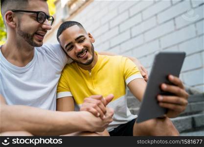 Portrait of happy gay couple spending time together and taking a selfie with digital tablet while sitting on stairs. Lgbt and love concept.