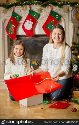 Portrait of happy girl and mother sitting at fireplace and wrapping Christmas presents