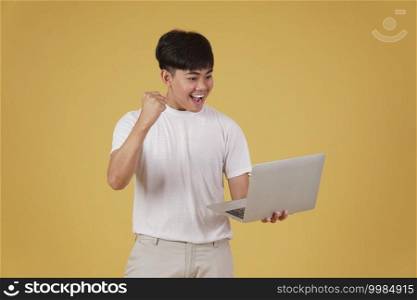 portrait of happy glad rejoicing cheerful young asian man dressed casually holding laptop computer doing fist winner gesture isolated on yellow studio background