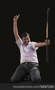 Portrait of happy Indian businessman with hockey stick pointing upwards while celebrating victory isolated over black background