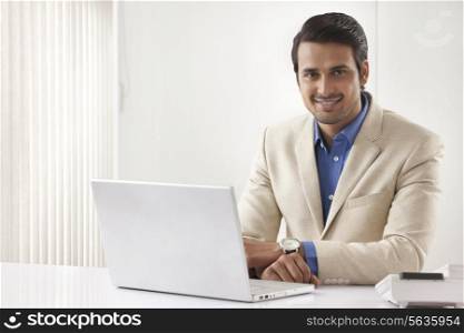 Portrait of happy Indian businessman with laptop sitting at office desk