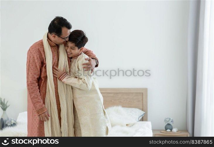 Portrait of happy Indian family standing and hugging in bedroom at home. Smart young father with little handsome son wear traditional dress, smiling and embracing each other, warm and cozy mood