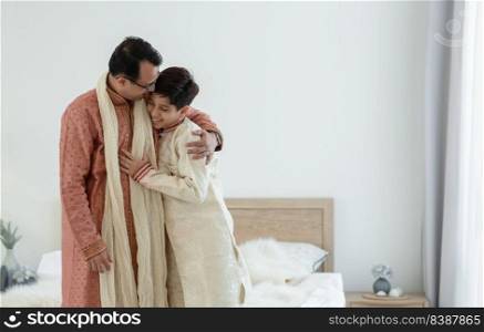 Portrait of happy Indian family standing and hugging in bedroom at home. Smart young father with little handsome son wear traditional dress, smiling and embracing each other, warm and cozy mood