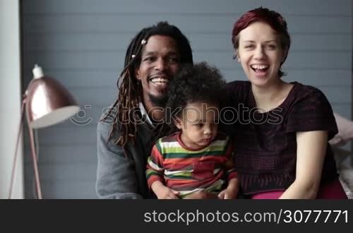 Portrait of happy interracial family with mixed race toddler son in the middle looking at the camera and smiling. Cheerful african american father with dreadlocks and caucasian mother cuddling their little curly pouty boy. Slow motion.