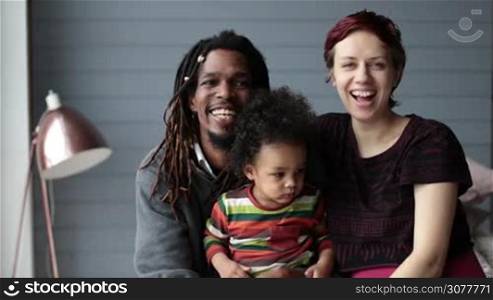 Portrait of happy interracial family with mixed race toddler son in the middle looking at the camera and smiling. Cheerful african american father with dreadlocks and caucasian mother cuddling their little curly pouty boy. Slow motion.