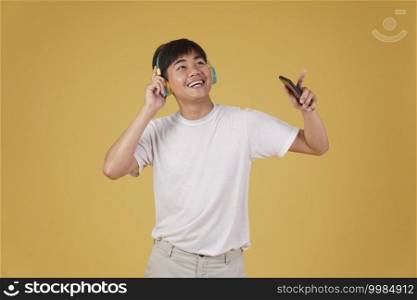 portrait of happy joyful cheerful young asian man wearing headphones listening to music on smartphone and dancing isolated on yellow studio background