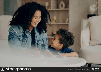 Portrait of happy joyful mixed race woman mom with long black curly hair sitting with little son in living room enjoying leisure time in each others company, learning how to draw. Happy maternity. Loving happy woman with long curly hair playing with little son at home