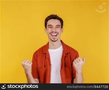 Portrait of happy joyful young man standing doing winner gesture clenching fists keeping isolated on yellow color wall background studio. Victory and success concept.