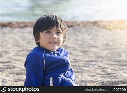 Portrait of happy kid boy smiling and looking up sitting on pebbles in the sunny day summer. Chid playing on beach in the weekend.