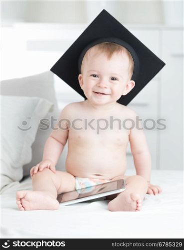 Portrait of happy laughing baby boy in graduation cap sitting with tablet pc