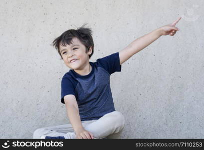 Portrait of happy little boy with smiling face pointing up, Kid with excited face sitting next to grey wall pointing finger, Child having fun playing outdoor, Positive children concept.