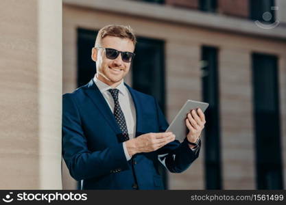 Portrait of happy male entrepreneur sends text message to colleague via touch pad, connected to wireless internet, updates software, wears trendy sunglasses and black suit. People and technology