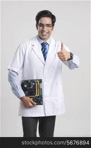 Portrait of happy male technician gesturing thumbs up while holding mother board over gray background