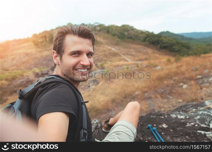 Portrait of Happy Man traveller sitting relaxing and taking selfie with smartphone in mountain view. Smiling Male Hiker enjoying with mobile phone on vacation, Holiday maker in nature.