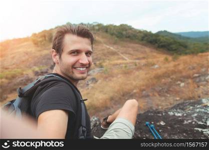 Portrait of Happy Man traveller sitting relaxing and taking selfie with smartphone in mountain view. Smiling Male Hiker enjoying with mobile phone on vacation, Holiday maker in nature.