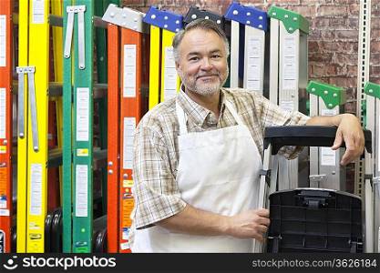 Portrait of happy mature store clerk standing by multicolored ladders in hardware shop