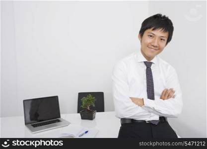 Portrait of happy mid adult businessman with arms crossed leaning on office desk