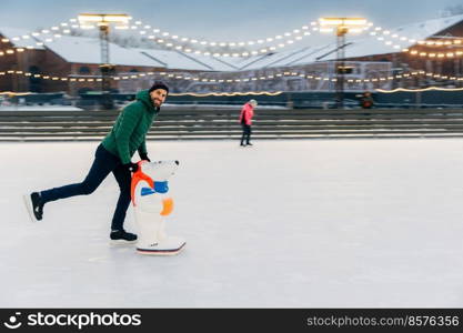 Portrait of happy middle aged male skater stands on ice ring, enjoys free time, has active lifestyle, being on ice arena. Smiling joyful man rejoices frosty winter weather. Skate aid. Hobby concept