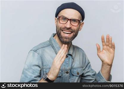 Portrait of happy middle aged man has fun indoor, raises hands, has pleased expression, wears glasses, hat and denim shirt, isolated over white background. People, positive emotions and feelings