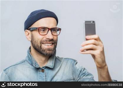 Portrait of happy middle aged smiling male wears denim jacket, hat and glasses, holds smart phone for making selfie, isolated over white background. People, technology and lifestyle concept.